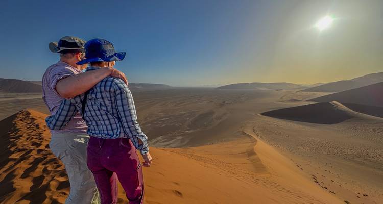 Paar, das auf einer hohen orangefarbenen Sanddüne in der Namib-Wüste steht und über ein weites Tal auf die helle Sonne blickt