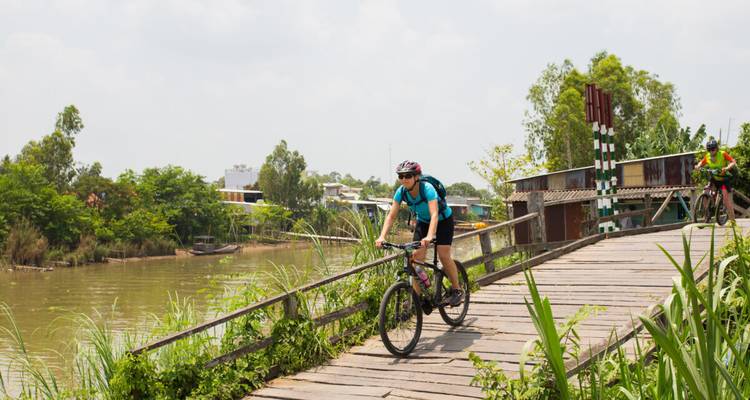 Radfahrer überqueren eine Holzbrücke über einen ruhigen Fluss in einer ländlichen Umgebung des Mekong-Deltas.