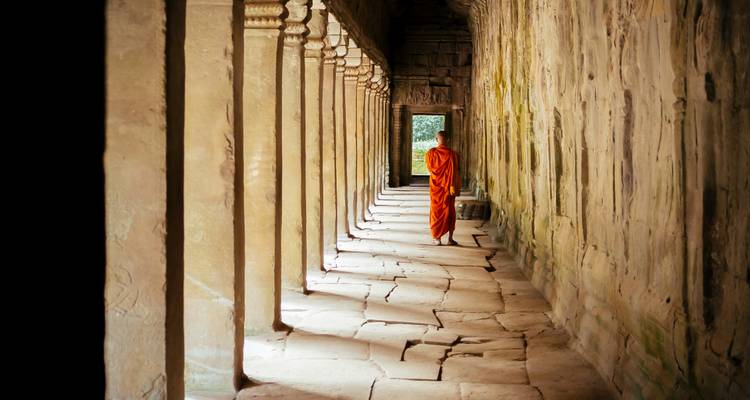Ein einzelner buddhistischer Mönch in orangefarbenen Roben geht durch eine steinerne Säulenhalle, die von sanftem Licht erleuchtet wird.