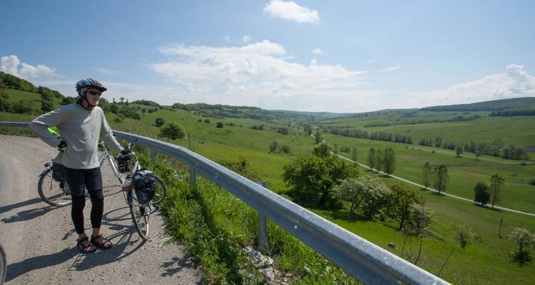 Ein Radfahrer pausiert neben einer Landstraße mit Blick auf sanfte grüne Hügel und Täler unter einem strahlend blauen Himmel.