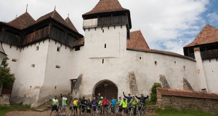 Eine große Gruppe von Radfahrern hebt ihre Hände vor einer weiß verputzten befestigten Kirche mit mittelalterlichen Türmen.