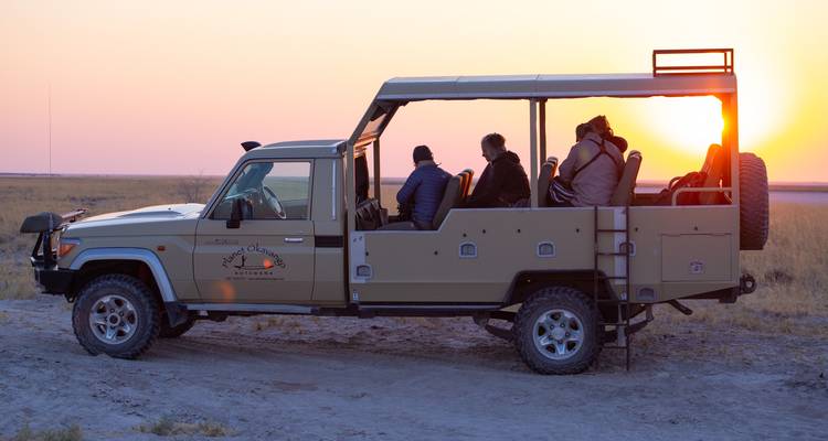 Offenes Safariauto mit Passagieren, die den Sonnenuntergang über trockenem Savannengelände beobachten.