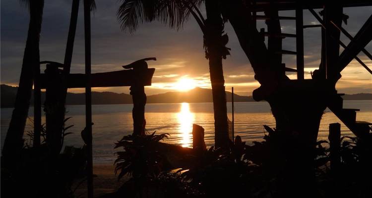 Un coucher de soleil tropical paisible se reflétant sur la mer, encadré par les silhouettes de palmiers et de structures en bois rustique sur la plage