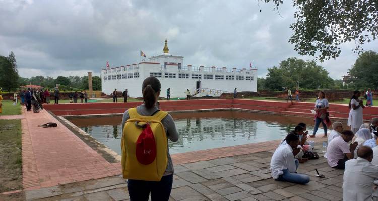 Un visiteur avec un sac à dos jaune observe l'étang sacré et le temple blanc Maya Devi tandis que des foules se rassemblent autour de l'eau.