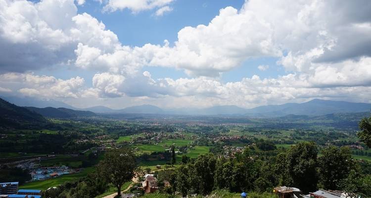 Vue panoramique d'une vallée luxuriante avec des habitations dispersées, entourée de collines et de montagnes sous un ciel nuageux.