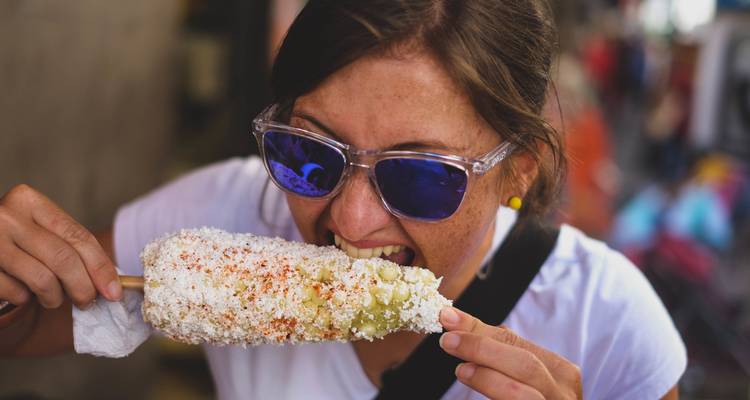 Traveller bites into a chilli-dusted elote street snack while wearing mirrored sunglasses.