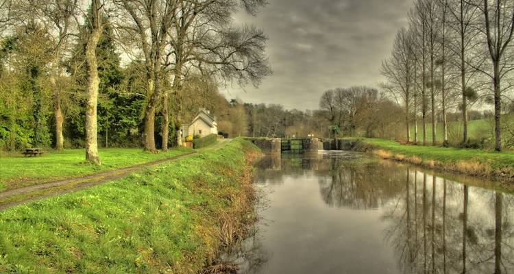 Ruhiger Kanal, umgeben von Bäumen und bewölktem Himmel, mit einer Schleuse in der Ferne.