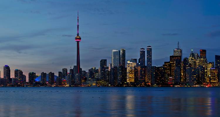 Evening panorama of Toronto skyline with CN Tower reflecting on calm lake waters at dusk.