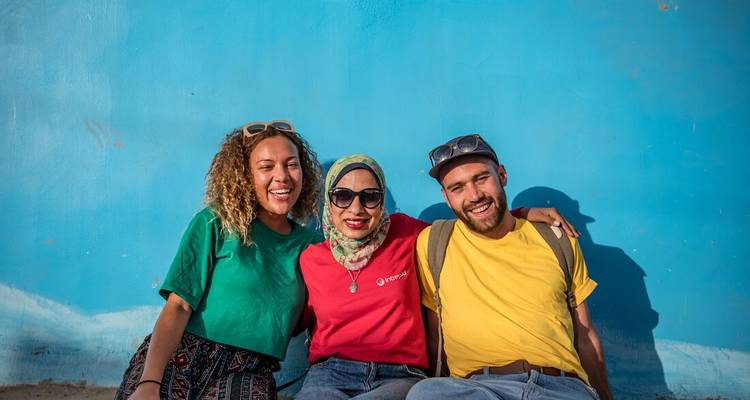 Three smiling travelers posing against a bright blue wall in sunlight