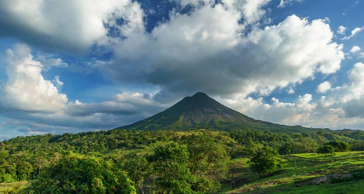 Arenal Volcano beneath dramatic clouds viewed across rolling green countryside