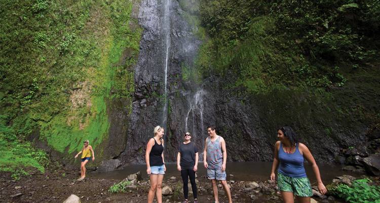 Travellers stand at the base of a tall jungle waterfall surrounded by lush green cliffs.
