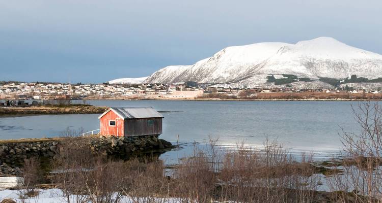Kleine rote Fischerhütte auf einem felsigen Steg mit Blick auf einen ruhigen Fjord, der von schneebedeckten Gipfeln und einem Küstendorf umgeben ist.