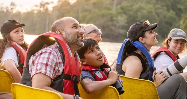 Groupe familial en gilets de sauvetage observant avec enthousiasme la faune sauvage depuis un petit bateau fluvial au coucher du soleil.