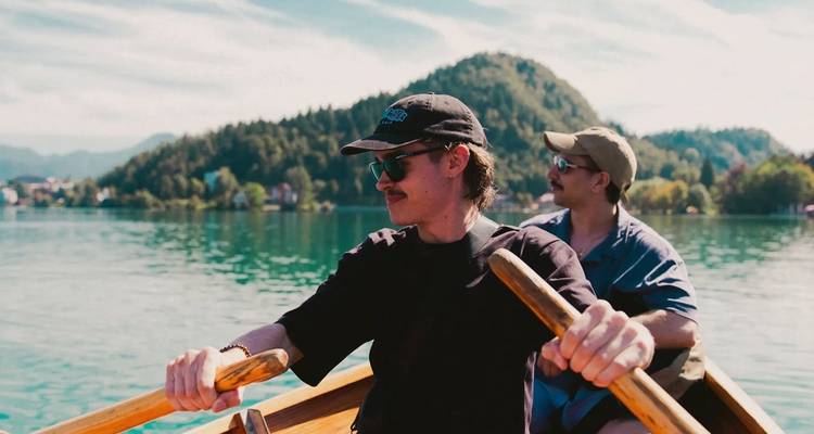 Two young men row a wooden boat across a tranquil turquoise lake surrounded by forested hills.