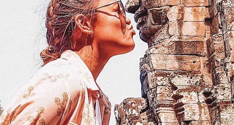 Mujer interactuando con humor con una escultura antigua de rostro de piedra.