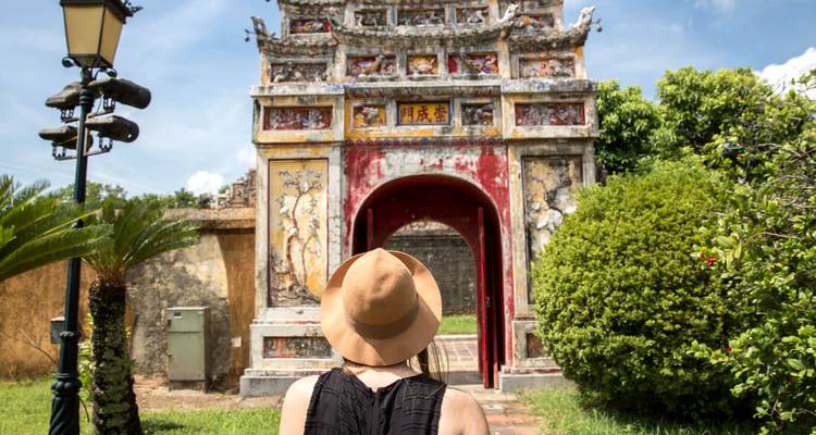 A female traveler wearing a hat gazes at an ornate weathered gateway in a historic Asian temple complex.