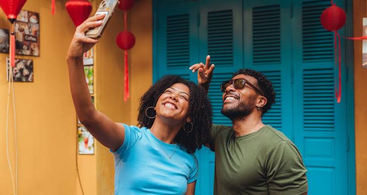 Un couple heureux prend un selfie devant une porte turquoise éclatante et des lanternes rouges à Hoi An.