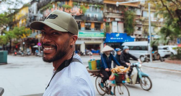Voyageur souriant dans une rue animée de Hanoï avec des scooters flous et des bâtiments coloniaux en arrière-plan.
