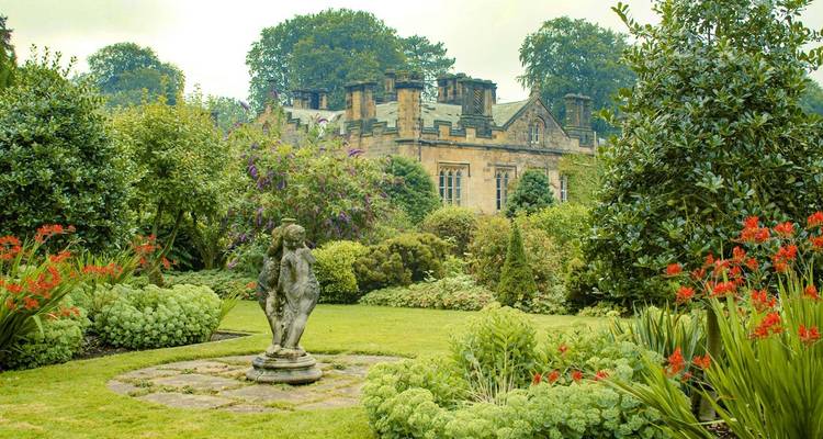 A serene garden with a historical manor and statues, lush with greenery and flowers.