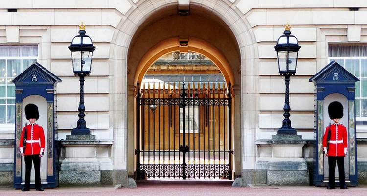 Dos guardias reales uniformados están de pie en casetas de vigilancia afuera de las puertas ornamentadas de un gran palacio.