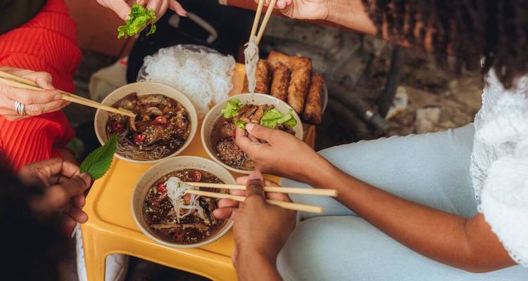 Vista aérea de comensales compartiendo tazones de caldo vietnamita con fideos y rollitos de primavera con palillos