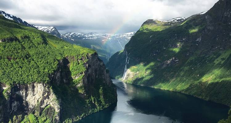Luftaufnahme eines tiefen norwegischen Fjords mit steilen grünen Felswänden, hohen Wasserfällen und einem zarten Regenbogen unter dramatischen Wolken.