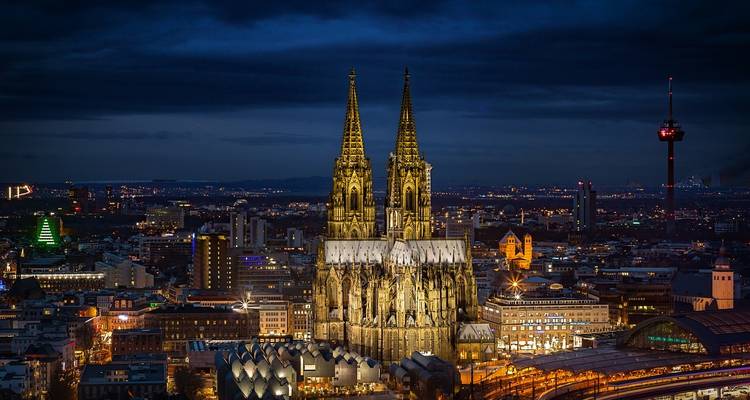 Una catedral gótica iluminada por la noche con las luces de la ciudad al fondo.