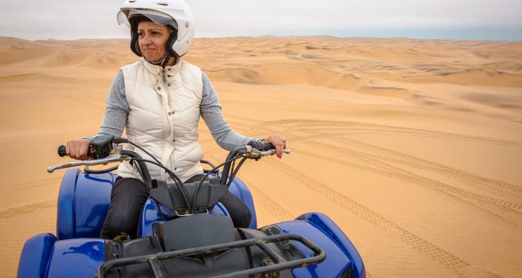 Mujer con casco monta un cuatriciclo azul a través de dunas de desierto ondulantes bajo un cielo nublado.