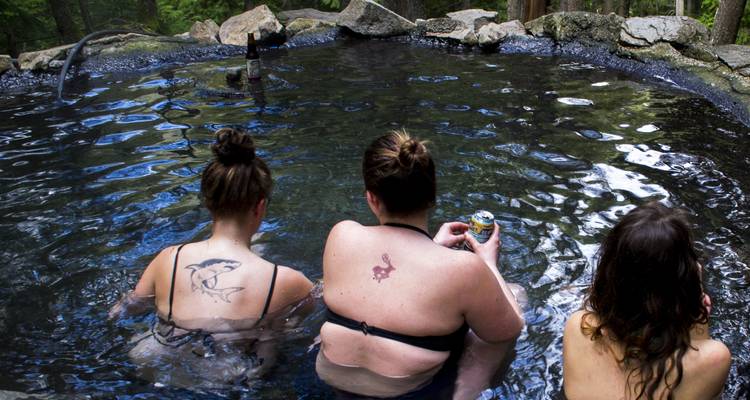 Trois femmes se détendent dans une source thermale naturelle en forêt, dégustant des boissons tout en étant immergées dans une eau minérale foncée.