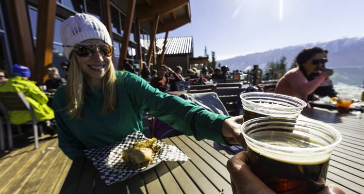 Des gens trinquent avec des chopes de bière sur une terrasse de montagne ensoleillée tout en savourant un déjeuner décontracté.