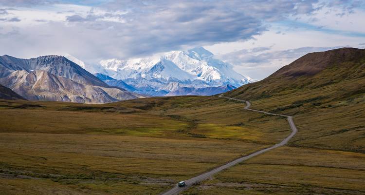 Une route sinueuse traverse la toundra dégagée en direction des pics enneigés du Denali sous des nuages spectaculaires