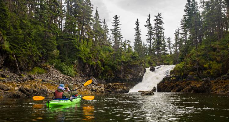 Un kayakiste solitaire pagaie en direction d'une cascade forestière entourée de falaises rocheuses