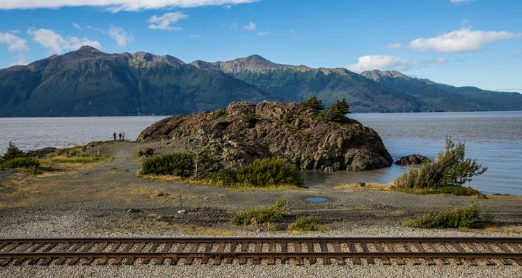 La péninsule rocheuse s'avance dans une large baie avec des voies ferrées au premier plan et un massif montagneux en arrière-plan