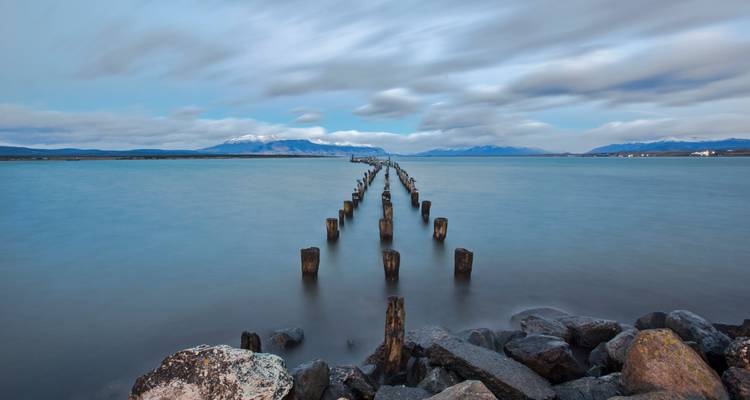 Des poteaux de vieux quai en bois menant à un fjord bleu calme avec des pics enneigés au loin au crépuscule