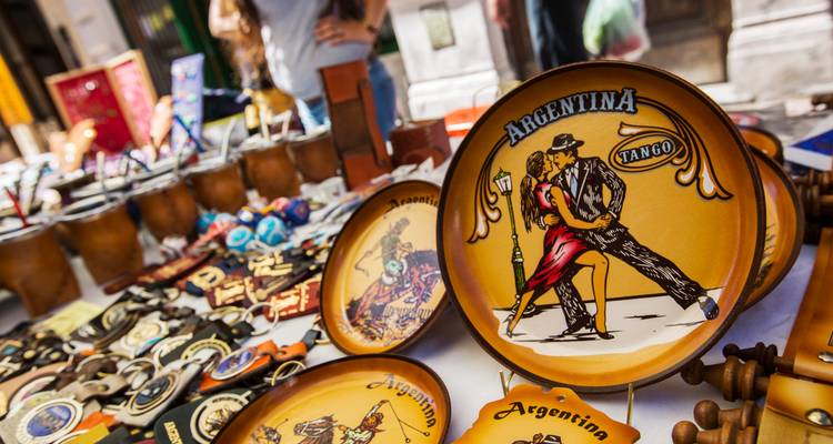 Des assiettes de souvenirs aux couleurs vives et thème tango sur un étal de marché avec des clients en arrière-plan.