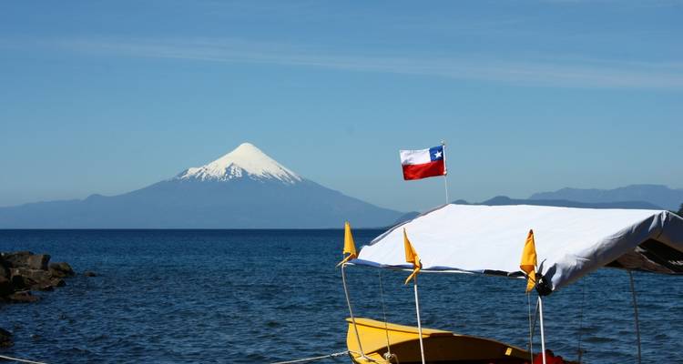 Vista lejana de un volcán nevado con una bandera y un lago en primer plano.