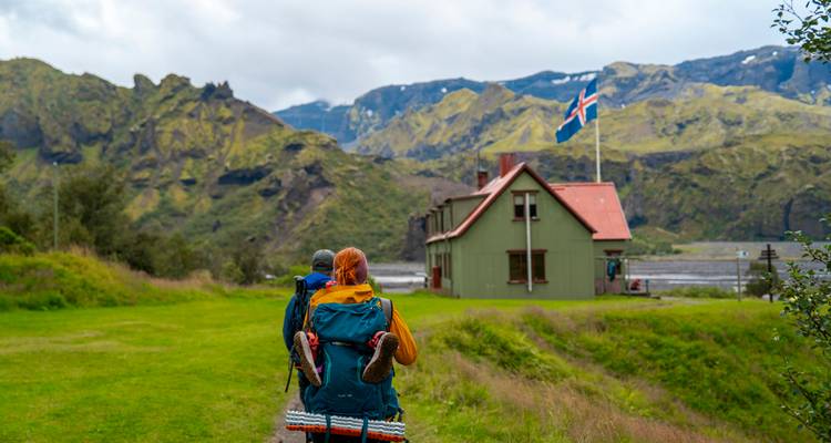 Deux routards se dirigent vers une cabane verte arborant le drapeau islandais au milieu de montagnes couvertes de mousse