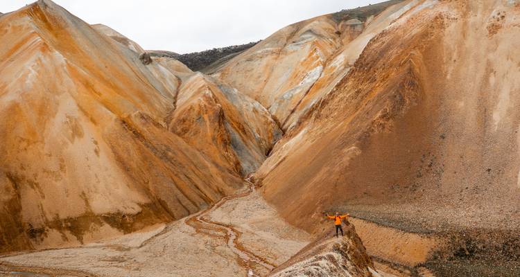 Randonneur en veste orange se tenant sur une crête de rhyolite acérée parmi les collines colorées de Landmannalaugar