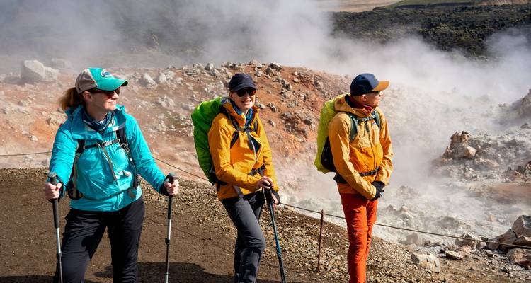 Tres excursionistas hacen una pausa cerca de respiraderos geotérmicos humeantes en una ladera volcánica escarpada.