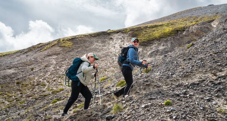 Deux randonneurs gravissent une pente de gravier meuble avec des bâtons de trekking sous un ciel partiellement nuageux.