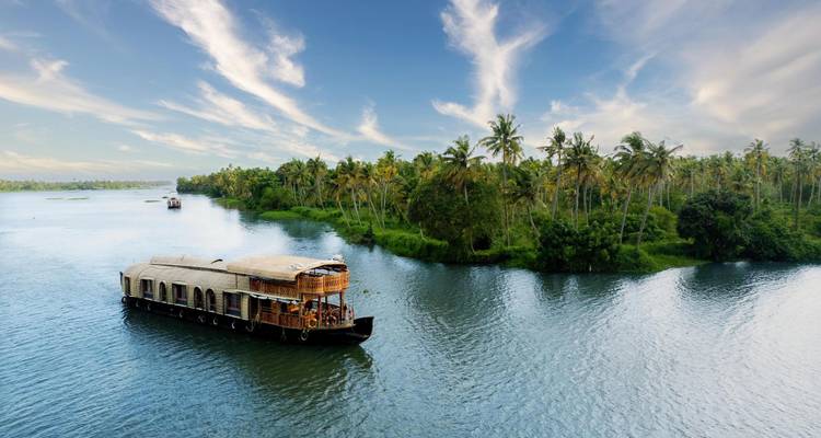 Traditional wooden houseboat cruises along Kerala backwaters framed by dense coconut palms and blue sky.