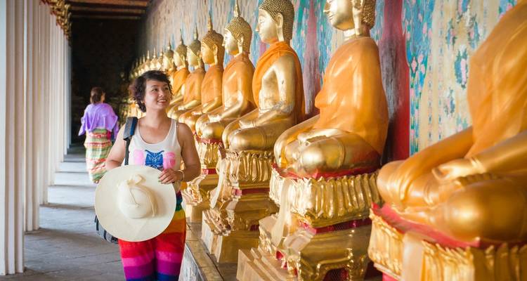 Una mujer sosteniendo un sombrero, caminando junto a una fila de estatuas doradas de Buda.