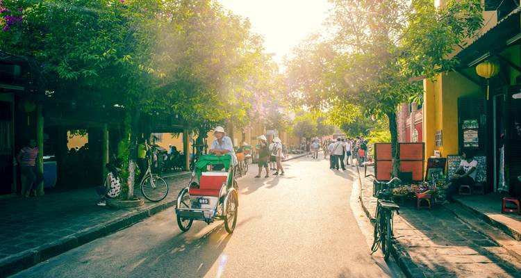 Escena callejera en Hoi An con bicicletas y una persona montando un rickshaw bajo un sol brillante.