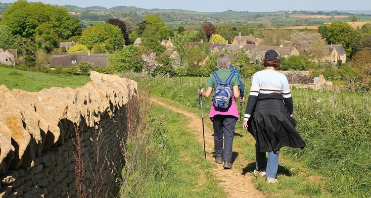 Dos personas caminando por un sendero en un paisaje rural con muros de piedra y cabañas.