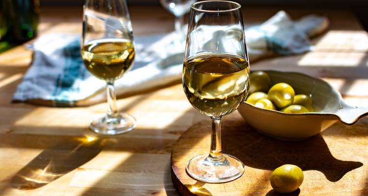 Sonnendurchflutete Gläser mit hellem Sherry-Wein und Oliven auf einem rustikalen Holztisch.