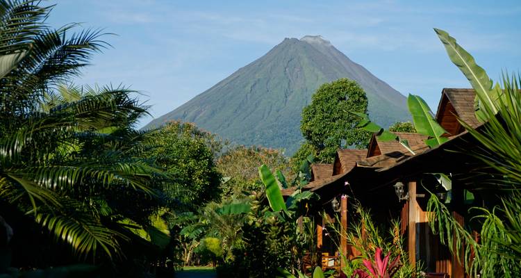 Volcan Arenal au loin, verdure luxuriante et cabanes au premier plan.
