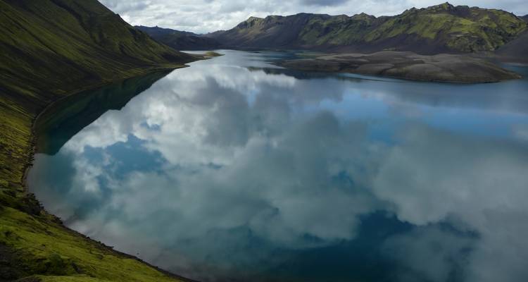 Lago azul espejo quieto arropado por empinadas laderas volcánicas verdes que reflejan dramáticos patrones de nubes
