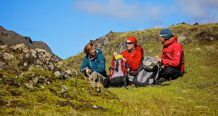 Tres excursionistas se sientan en el pasto con mochilas, compartiendo un refrigerio con un fondo de montañas y cielo azul