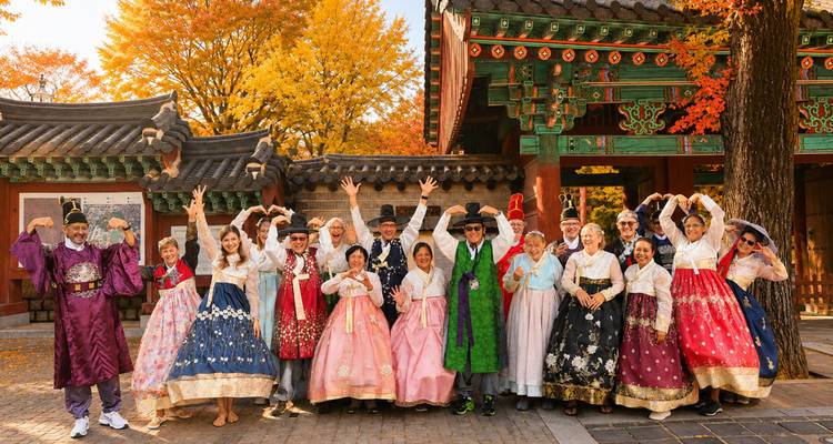 Un grand groupe vêtu de hanbok aux couleurs éclatantes célébrant devant une cour de palais coréen traditionnel avec un feuillage automnal.