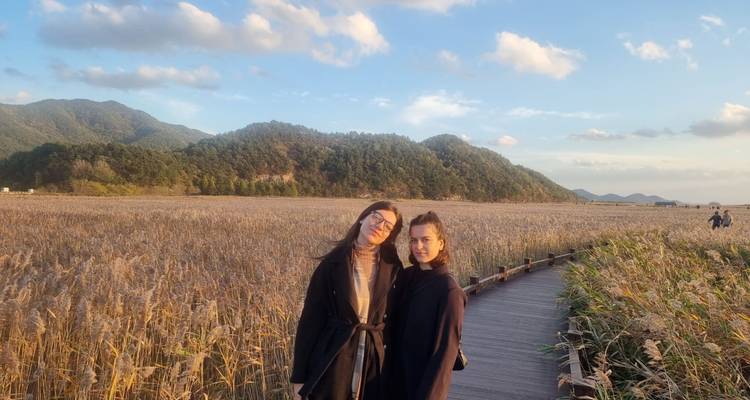 Deux femmes se tenant debout sur une passerelle en bois traversant de vastes champs de roseaux sous un ciel de soirée aux teintes pastel.